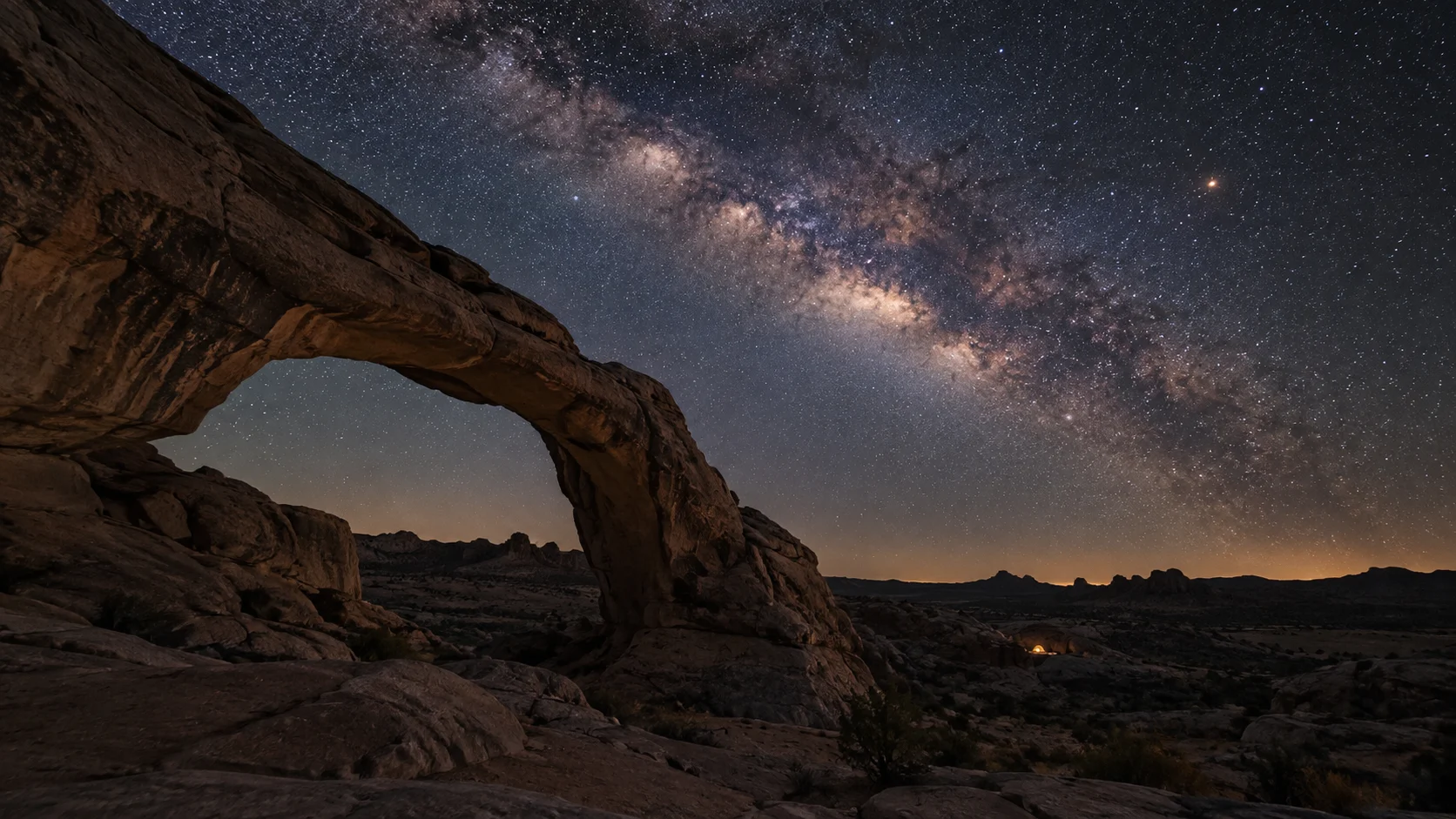 The Milky Way glows above a natural stone arch in the desert.
