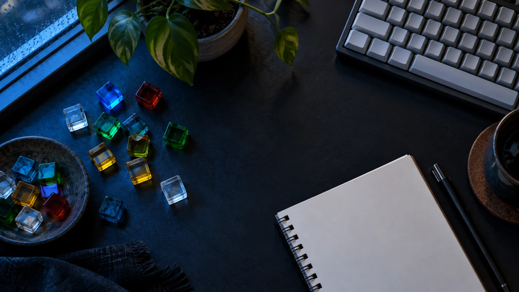 Colored glass tiles, a plain keyboard, blank notebook, and plant cover a dark desk.