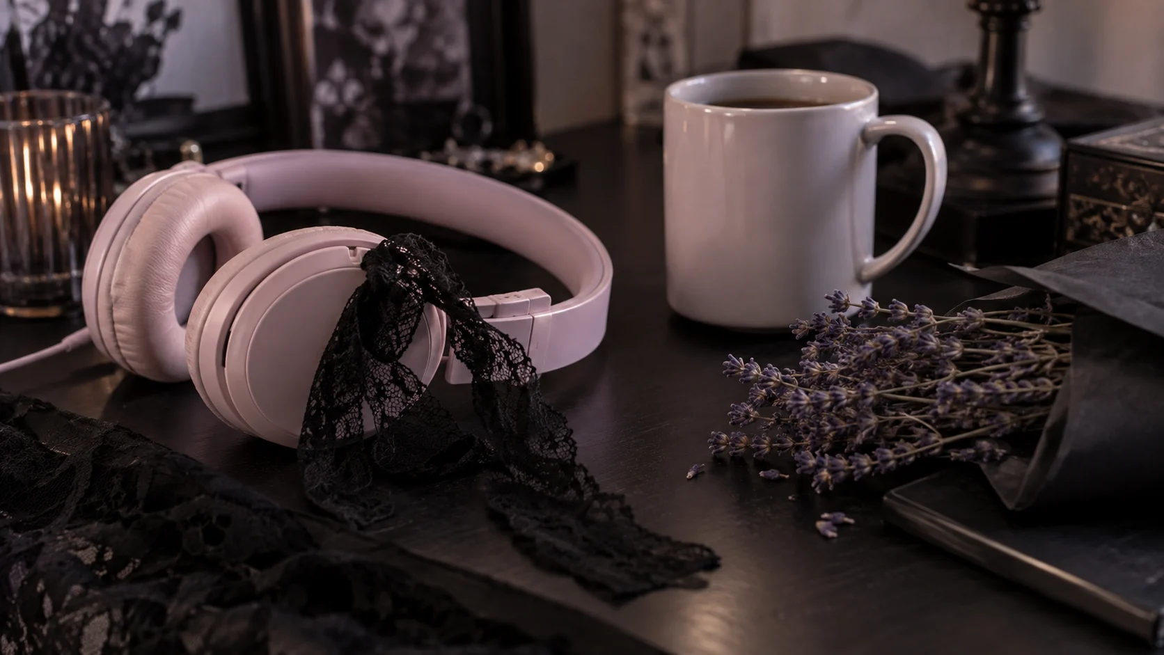 Pale pink headphones, black lace, and lavender flowers sit on a dark desk.